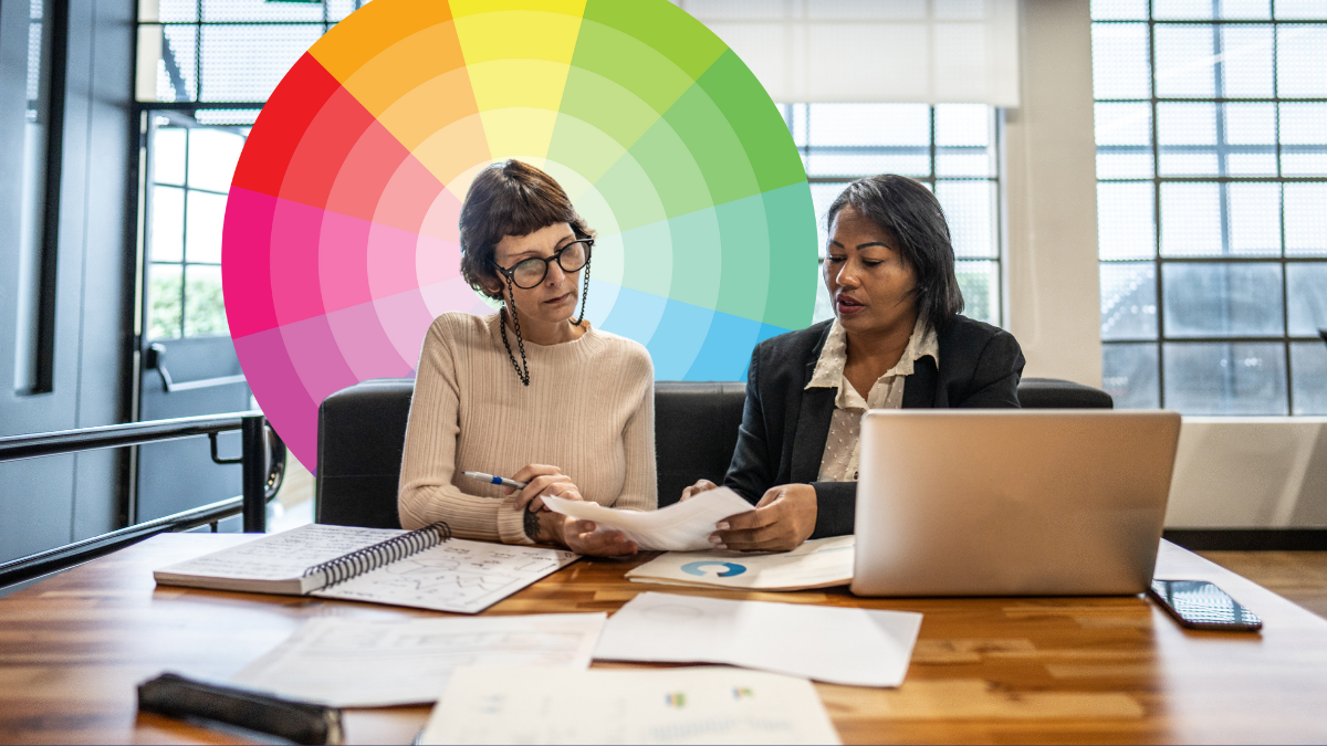 Two female business leaders sitting at desk looking at reports colourful wheel behind one leader respresenting the feelings wheel
