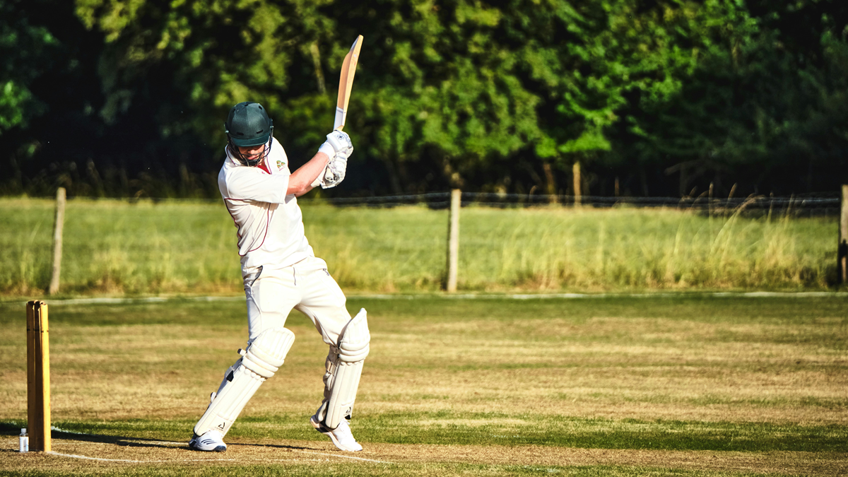 Cricket batsman in whites and pads playing a shot on a grass pitch, bat raised, poised and composed at the crease