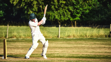 Cricket batsman in whites and pads playing a shot on a grass pitch, bat raised, poised and composed at the crease