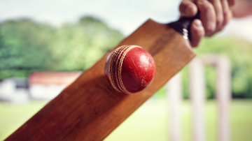 Worn red leather cricket ball meeting the middle of a wooden bat in front of the stumps, backlit on a sunny ground